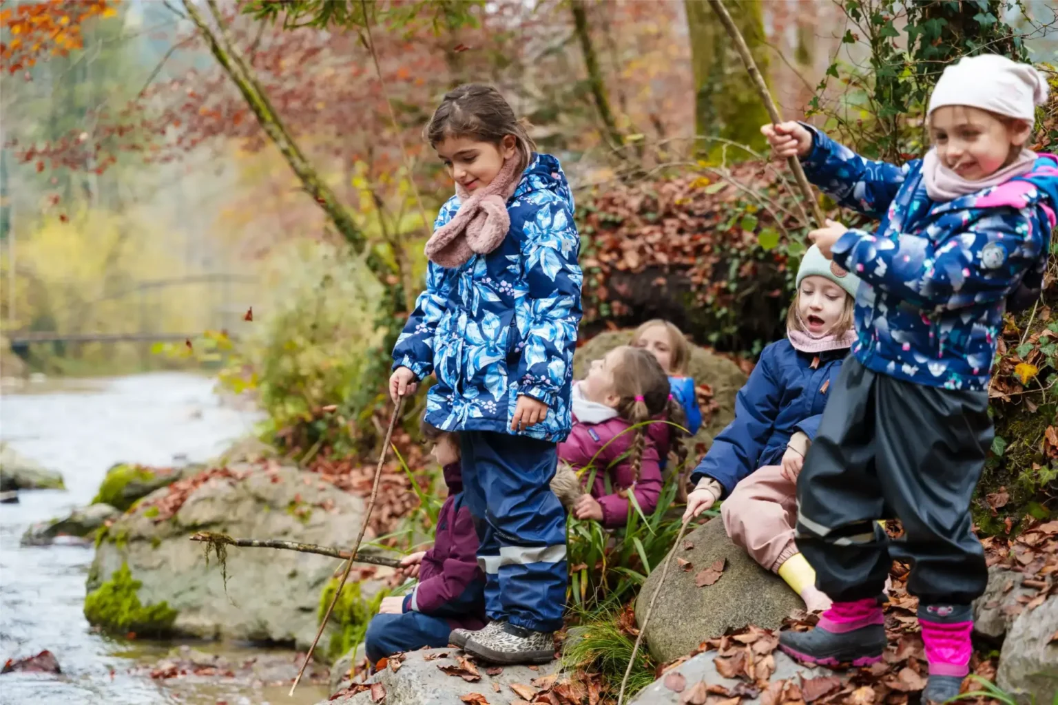A group of young children in colorful jackets sit and stand on rocks by a creek in a forest, holding sticks and playing near the water on an autumn day.