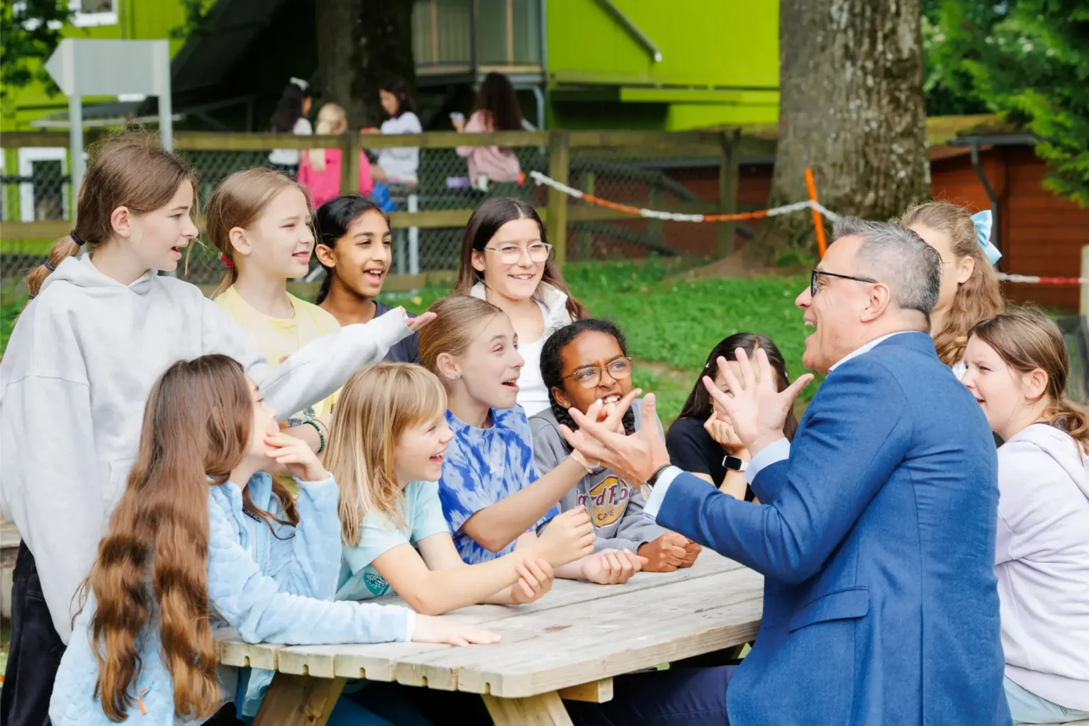 A group of smiling children sit around a wooden picnic table outdoors, excitedly engaging with an adult man in a blue suit who gestures animatedly with his hands. Trees and a green building are in the background.