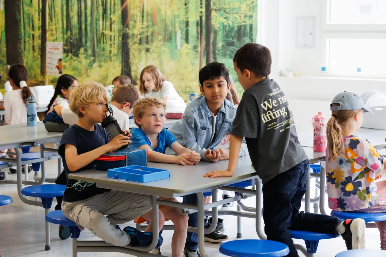 A group of young children sit and talk at a cafeteria table in a bright room with a forest mural on the wall. Some children are eating, drinking, or chatting, while others sit at nearby tables.
