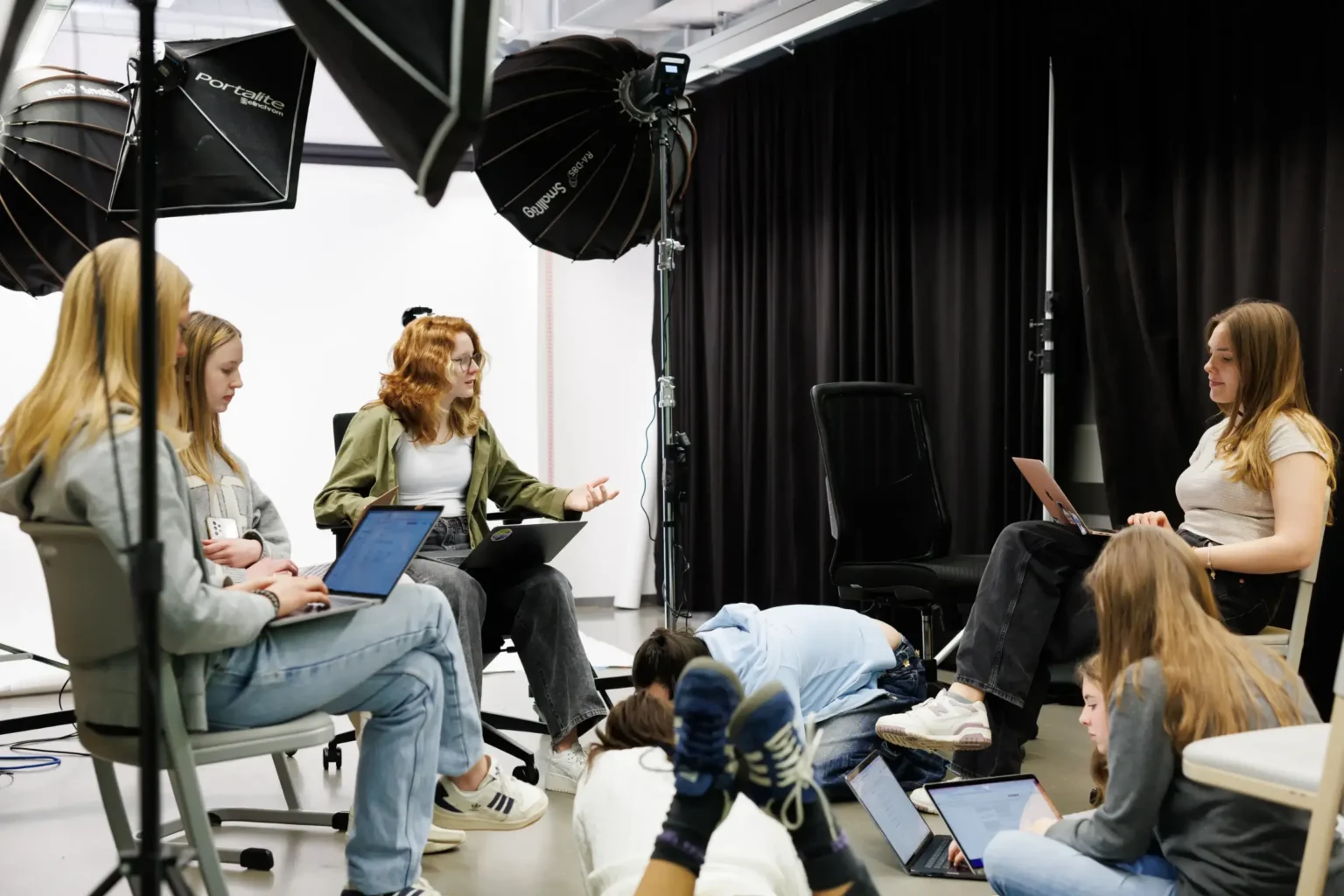 A group of young people work on laptops in a studio with lighting equipment and black curtains. Some sit on chairs, others on the floor, appearing engaged in discussion or collaboration.